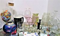 Overview of glassware lot showing six glass knobs, pink footed bowl, various glasses, and brown stoneware creamer.