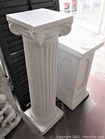 Tall white plaster fluted column pedestal with Ionic style capital and square base alongside shorter white paneled rectangular pedestal.