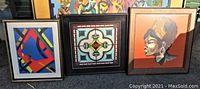 Photo showing all three framed needlework art pieces set on ground side by side, featuring one tribal lady portrait and two abstract geometric designs.