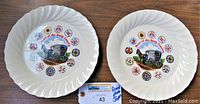 Two white ceramic plates with a fluted edge, decorated with Amish buggy and quilt designs, placed side by side on a wooden surface, showing both plates at once.
