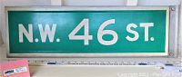 Full view of green and white 'N.W. 46 ST' aluminum street sign with hanger bracket attached, placed flat on a white surface with a ruler for scale.