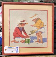 Full view of the framed needlepoint artwork showing two children playing on a beach with sand bucket and spade.