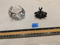 Photo showing both the diamond-shaped glass paperweight and the small metal pill case with a chicken figure on top, placed alongside a ruler for size reference.