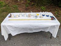 Wide view of a large table covered with a white cloth displaying a variety of vintage glassware including cups, bowls, mugs, wooden holder for small glasses, and other small glass items arranged in rows.