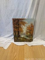 Full view of oil painting showing fall landscape with trees and river against a light blue sky on a white fabric background.