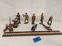 Ten old lead toy soldiers in various poses including standing, kneeling with binoculars, operating a machine gun, lying prone with rifle. Brown painted figures with green bases and silver helmets. Shown with ruler for scale about 3 inches tall.