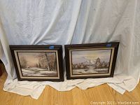 Pair of framed oil on canvas mountain scene paintings set against a white fabric background