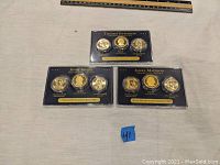 Three lucite collector cases each holding three presidential dollar coins representing Thomas Jefferson, John Adams, James Madison, and others, arranged on a table with a ruler above for size reference.