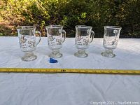 Four vintage Anchor Hocking footed coffee mugs with glass handles and varied coffee beverage prints in white and gold, displayed outdoors on a white cloth with measuring tape.