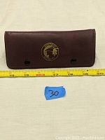 Brown vintage darts case closed, placed next to a tape measure showing approximately 7.5 inches width. The front features a gold logo of a dart player.