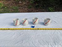 Four white ceramic coffee mugs with red stripe and gold TWA logo placed in a row on a white cloth with measuring tape showing size.