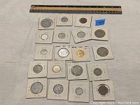 Overview photo showing 23 varying world coins in white cardboard holders with staples, different sizes and metals including silver and gold tones.