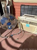 Photo of Fedders window AC unit and Patton high velocity fan placed outside against a wall, showing front view of AC unit and side view of fan.
