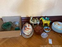 Shows ceramic camel figurine, ceramic monkey riding a horse figurine, glass dish with bunny lid, dinner plates, brown ovenware bean pot, and other ceramic and glass items
