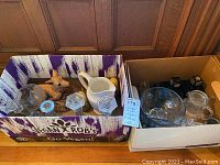 Overview of glass and decorative items in two boxes, including white ceramic pitcher, wooden sculptures, glass lidded jars, candlesticks, decanters, and salt and pepper shakers.