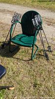 Green metal garden chair with ribbed backrest on grass, six black metal torches with ribbed glass jars leaning on chair, attached clamps for deck railing mounting visible
