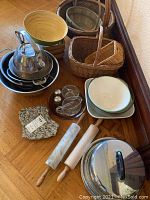 Overview of the lot showing stainless steel kettle, mixing bowls, metal strainer, salt and pepper shakers, glass cups with saucers, ceramic plates, wooden rolling pins, marble trivet, and wicker baskets.