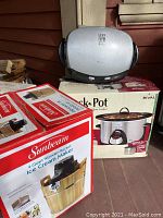 Photo of three boxed kitchen appliances placed on porch: Sunbeam 4 Quart Wooden Bucket Ice Cream Maker, Rival Crock-Pot slow cooker, and George Foreman grill.
