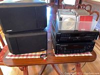 Two black Bose speakers stacked with Sony stereo receiver and CD player on a table with red and white checkered cloth, connected with cables.