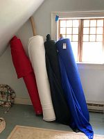 Four large bolts of fleece fabric in red, off-white, black, and blue resting upright against a wall by a window.
