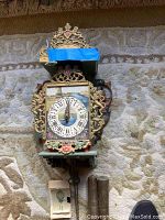 Friesian Chair wall clock with metal adornment and floral hand painting, clock face with Roman numerals, two metal weights, and visible chains on carpeted floor.