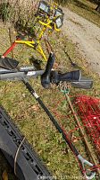 Image showing yellow dual-head contractor task light on stand, black rubber boots, long-handled garden tools including rakes and snow shovels, and red tomato cages on grass.