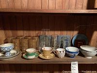 Clear glasses with ornate gold or brownish pattern along with brown woven vases and Bone China cups with saucers in front of a wood panel wall.