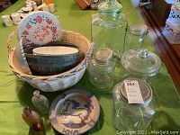 View of all items: white wicker basket with ceramic bowls and plates, several glass jars, and vintage bottles on a green tablecloth.