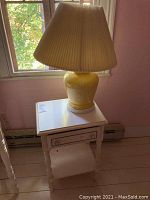 Photo of the yellow ceramic lamp with pleated shade placed on the white Bassett side table next to a window.