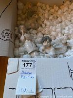 Box containing the three Lladro figurines surrounded by packing peanuts. Visible are parts of the figurines showing delicate features and soft pastel coloring.