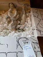 Three Lladro figurines inside a box with packing peanuts, including the two seated angels and the woman with geese