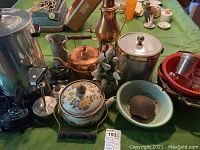 Several kitchen items including West Bend coffee percolator, copper kettle, ice bucket, enamel items, and pewter mug arranged on a table with a green table cloth.
