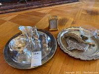 Two silver plate serving trays, one holding a pair of salt and pepper shakers, a creamer, a sugar bowl, and the other holding a crumb sweeper and the brass Tiffany clock.