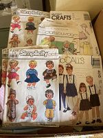Photo of multiple vintage sewing pattern packets for doll and baby clothes including Simplicity and McCall's Crafts brands, overlapping each other on fabric surface.