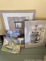 Photo showing multiple pieces of wall art stacked together including framed and unframed prints and paintings. Visible signed painting of animals and outdoor scenes, with some artworks in protective plastic sleeves.