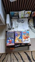 Photo showing Nintendo Wii console on stand, two white Wii remotes with cables, sensor bar stands, and assorted DVDs and VHS tapes.