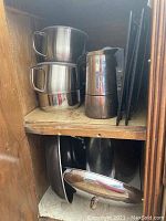 Three stacked stainless steel sauce pans, a metal carafe, multiple black spiral slicers, and a large stock pot with lid visible on a wooden shelf