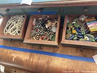 Three wooden drawers filled with assorted costume jewelry including pearl-like bead necklaces, mixed metal and bead jewelry, and colorful bracelets. Drawers are open on a wooden surface.