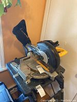 Side view of Dewalt compound miter saw on black stand next to window showing the base with angle measurements and dust collection bag attached.