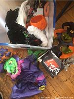 Full view of two bins containing Halloween decorations including a plastic orange pumpkin, various fabrics, and witch-themed items, with scattered decor on a wooden floor.
