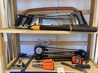 Photo showing multiple handsaws including tree saw, finish saw, coping saw, two hack saws, Fiskar's trimmer, Vemco drafting arm, two combination squares, three chisels, and other measuring and hand tools.