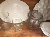 White ceramic turkey imprint platter, egg platter, covered glass jar, clear glass vase and small glass containers on wooden surface.