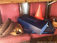 Photo of a worn Rawlings child-sized catcher mitt, NYCFC boxed kits and scarf, and an orange traffic cone on a red patterned seat under a wooden table.