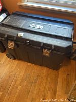 Front view of black Stanley tool storage box with wheels and metal latches on hardwood floor.