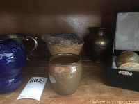 Shelf view of six decor pieces including blue ceramic vase, silver plate vessel, beige bowl, small ceramic pot, two wine goblets, and boxed eggs