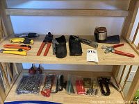 Photo of shelf with assorted hand tools including box cutters, grommet punch pliers, multi-tool, drill bits, hex keys, and industrial staple gun