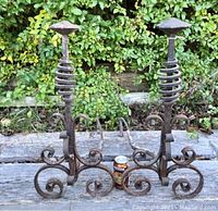 Pair of antique hand-wrought iron andirons outdoors on weathered wood surface with greenery background, showing full front profile of both pieces with visible spiraled irons curling in opposite directions, scroll feet, and square hammered tops.