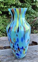 Full view of tall blue mid century modern glass vase with multicolored applied splash pattern in an outdoor setting.