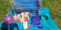 Full view of cage and all accessories arranged on blue cloth outdoors, items include cage with extra mesh, exercise ball, running wheel, food bags, litter box, feeding bowl, hut, scoop, and brush.