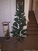 Photo of artificial green Christmas tree, approximately 6 feet tall, set up with metal stand and its original box visible.